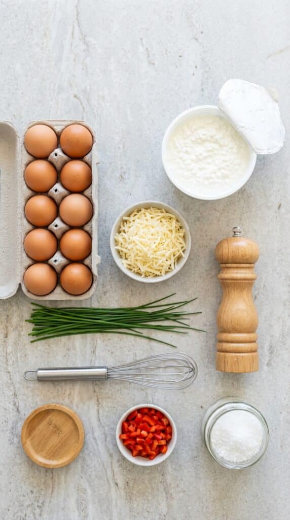 A flat lay overhead view showing brown eggs, a tub of cottage cheese, shredded cheese, and fresh chives on a stone counter.