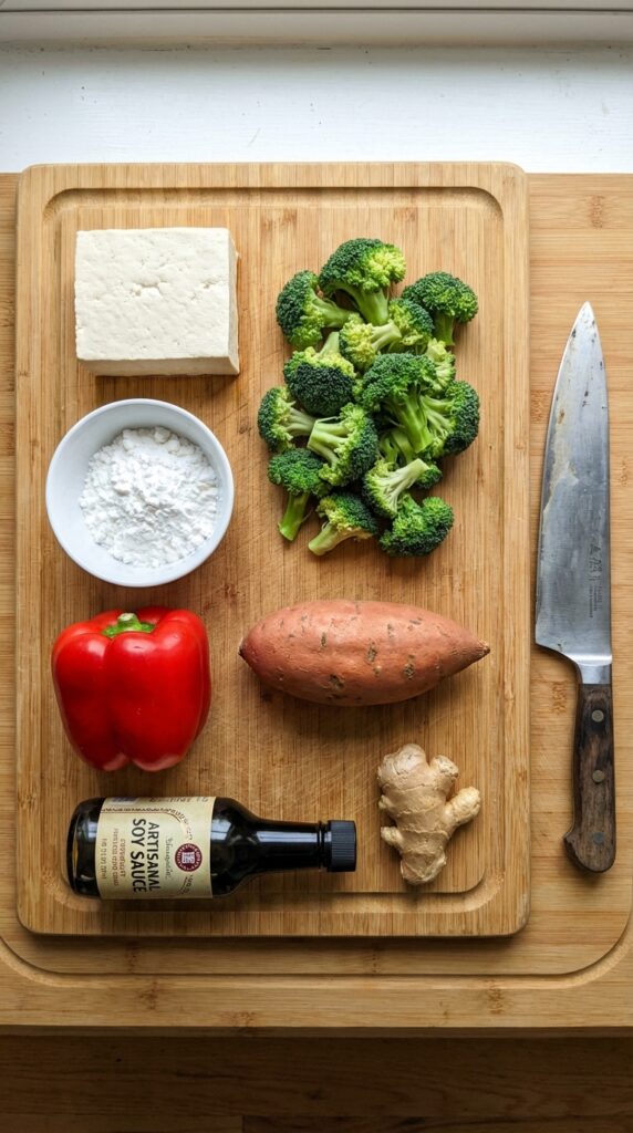 A flat lay showing a block of firm tofu, broccoli, red pepper, sweet potato, soy sauce, and cornstarch on a cutting board.