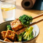 A close-up of chopsticks lifting a piece of glazed crispy tofu and broccoli from a rice bow