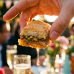 A close-up of a hand holding a single sticky baklava bite with honey dripping off.
