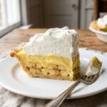 A close-up cross-section of a slice of banana cream pie on a plate, showing distinct layers of crust, bananas, custard, and whipped cream.