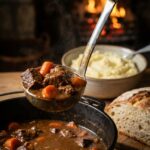 A close-up of a ladle lifting thick beef stew with vegetables, with mashed potatoes in the background.