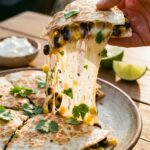 A close-up of a hand lifting a quesadilla wedge with a long cheese pull, dipping it into sour cream.