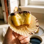 A close-up of a hand holding a Boston cream cupcake with a bite taken out, showing the vanilla cream filling oozing out.