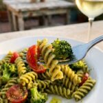 A close-up of a fork lifting a bite of green pesto pasta with a cherry tomato, with a sunny patio in the background