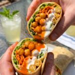 A buffalo chickpea wrap cut in half showing spicy orange chickpeas, lettuce, avocado, and ranch dressing inside.