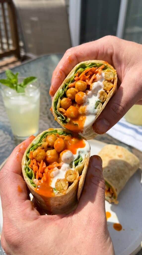 A buffalo chickpea wrap cut in half showing spicy orange chickpeas, lettuce, avocado, and ranch dressing inside.