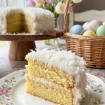 A close-up of a slice of coconut cake on a floral plate with Easter eggs in the background.