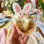 A close-up of a hand holding a bunny cupcake with the wrapper peeled down, showing the soft vanilla cake inside.