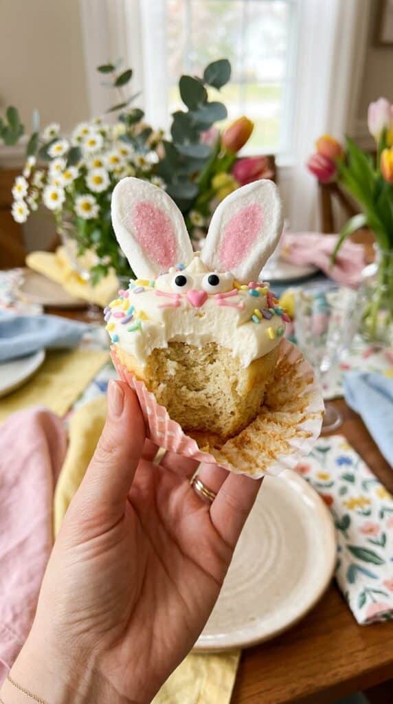 A close-up of a hand holding a bunny cupcake with the wrapper peeled down, showing the soft vanilla cake inside.