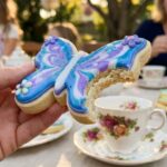 A hand holding a butterfly cookie with a bite taken out of it, set against a garden background.