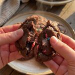 A close-up of hands breaking a soft sprinkle cookie in half, showing a gooey, cake-like interior.