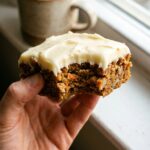 A close-up of a hand holding a bitten carrot cake bar, showing the moist texture and thick frosting.