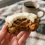A close-up of a hand holding a bitten carrot cake cookie, showing the soft inside texture.