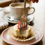 A close-up of a fork cutting into a mini cheesecake cupcake on a plate, showing the creamy texture.