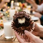 A hand holding a unwrapped chocolate cupcake with a bite taken out, showing the cheesecake filling inside.