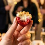 A close-up of a hand holding a bitten cheesecake-stuffed strawberry, showing the filling inside.