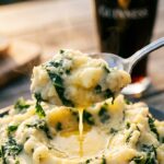 A close-up of a spoon lifting buttery mashed potatoes with kale, with a beer in the background.