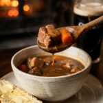 A close-up of a spoon lifting a bite of pork stew from a bowl, with soda bread on the side.