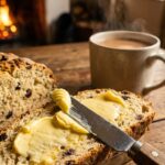 A close-up of a knife spreading butter on a slice of soda bread with currants.