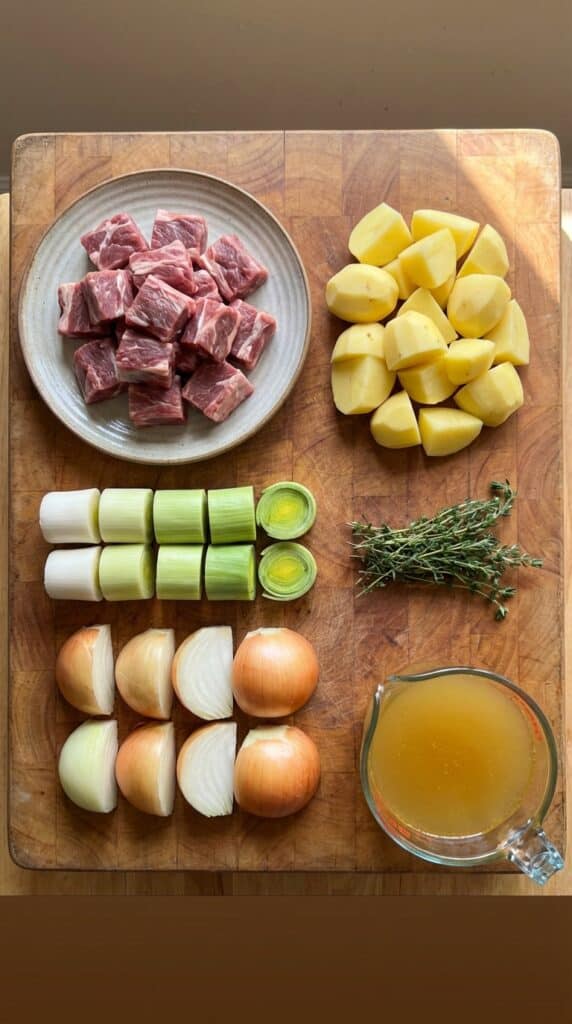 A flat lay showing raw lamb chunks, potatoes, leeks, onions, and herbs on a wooden board.