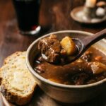 A close-up of a spoon lifting lamb and potato from a bowl of stew, with soda bread on the side.