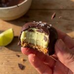 A close-up of a hand holding a chocolate-covered key lime pie bite with a bite taken out, showing the crisp shell and creamy filling.