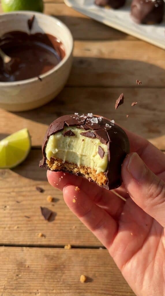 A close-up of a hand holding a chocolate-covered key lime pie bite with a bite taken out, showing the crisp shell and creamy filling.