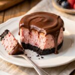 A close-up cross-section of a pink strawberry mini cheesecake with an Oreo crust and chocolate ganache, with a fork taking a bite.