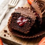 A close-up of a chocolate cupcake cut in half, revealing a bright red strawberry jam filling inside.