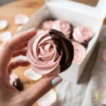 A hand holding a single pink meringue rose dipped in chocolate, with a gift box in the background.