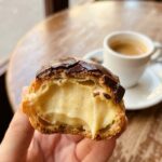 A close-up of a hand holding a bitten chocolate eclair, showing the airy pastry shell filled with vanilla cream.