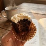 A close-up of a hand holding a half-unwrapped coffee cupcake with a bite taken out, showing the moist crumb and thick frosting.