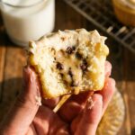A close-up of a hand holding a cupcake with a bite taken out, showing the raw cookie dough center.
