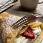 A close-up of a fork slicing through a strawberry crepe revealing the cream filling.