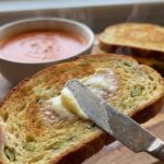 A close-up of a knife spreading butter on a warm, toasted slice of pickle bread.