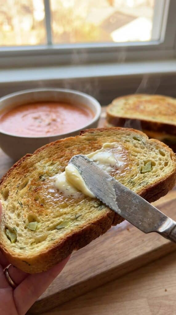 A close-up of a knife spreading butter on a warm, toasted slice of pickle bread.