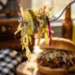 A close-up of a fork lifting creamy pickle coleslaw with a sandwich in the background.