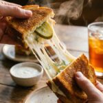 A close-up of hands pulling apart a grilled cheese sandwich to reveal stringy melted cheese and pickles.