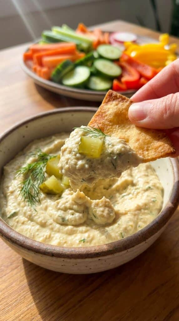 A close-up of a hand dipping a pita chip into creamy dill pickle hummus.