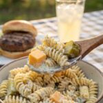 A close-up of a spoon lifting creamy pasta salad with a piece of pickle and cheese.