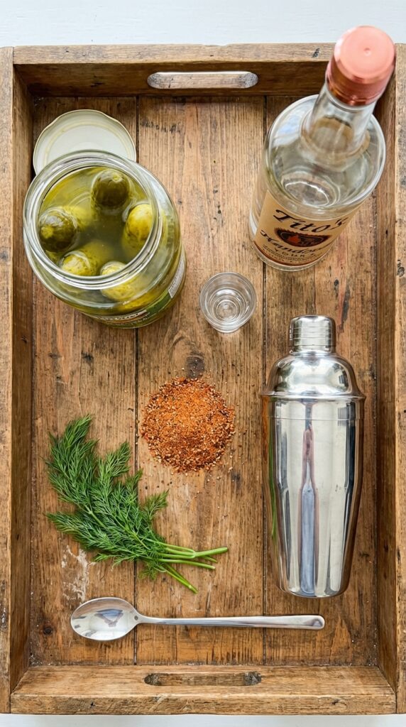 A flat lay showing a jar of pickles, vodka bottle, cocktail shaker, and red seasoning on a wooden surface.