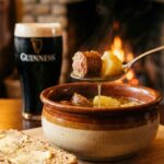 A close-up of a spoon lifting sausage and potato from a bowl of coddle, with soda bread on the side.