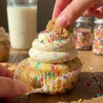 A close-up of a hand peeling the wrapper off a funfetti cupcake while dipping a graham cookie into the speckled frosting.