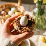 A close-up of a hand holding a chocolate spoon topped with a coconut nest and mini eggs.
