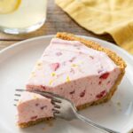 A close-up of a slice of frozen strawberry lemonade pie on a plate, showing the creamy texture and berry chunks.