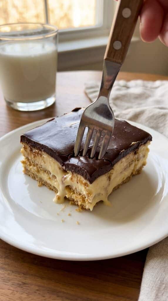 A close-up of a fork cutting into a soft slice of layered eclair cake on a plate.