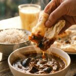 A close-up of a hand dipping naan bread into a bowl of hot eggplant curry.