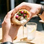 A close-up of two hands holding a loaded falafel wrap with tahini sauce dripping, ready to take a bite.