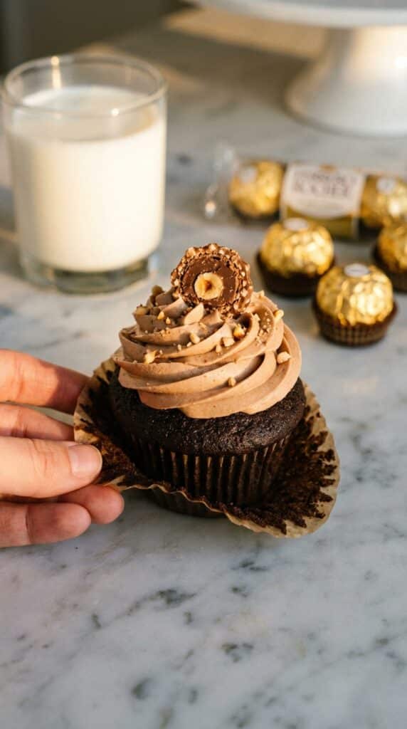 A close-up of a hand peeling the paper wrapper off a frosted chocolate hazelnut cupcake